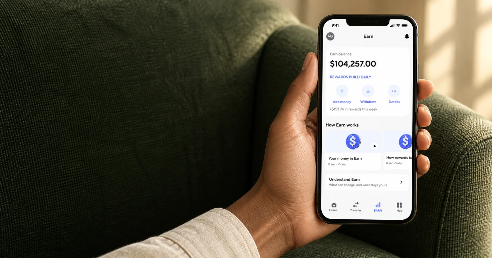 A person holding a phone showing a funded Dara Earn account, with an Earn balance of $104,257.00 and rewards this week of $153.74, beside a green woven armchair.